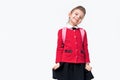 Adorable little girl in red school jacket, black dress, backpack shyly clings to a skirt and smile while posing on white