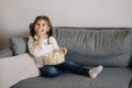 Adorable little girl sits on sofa and watching TV at home. Cute girl eating popcorn. Holiday mood Royalty Free Stock Photo