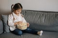 Adorable little girl sits on sofa and watching TV at home. Cute girl eating popcorn. Holiday mood Royalty Free Stock Photo