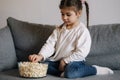 Adorable little girl sits on sofa and watching TV at home. Cute girl eating popcorn. Holiday mood Royalty Free Stock Photo