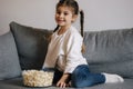 Adorable little girl sits on sofa and watching TV at home. Cute girl eating popcorn. Holiday mood Royalty Free Stock Photo