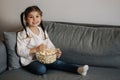 Adorable little girl sits on sofa and watching TV at home. Cute girl eating popcorn. Holiday mood Royalty Free Stock Photo