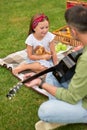 Adorable little girl sitting on a green grass in park and looking at her dad playing guitar