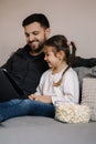 Adorable little girl watching cartoons on table with her father. Daughter with dad eating popcorn and laugh. Home mood Royalty Free Stock Photo