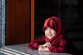 Adorable little muslim girl in traditional clothing, red hijab or niqab and red abaya smiling and watching out the window. Prawet