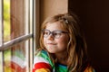 Adorable little preschool girl with eyeglasses sitting by the window. Thoughtful child looking out. Lonely kid. Royalty Free Stock Photo