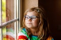 Adorable little preschool girl with eyeglasses sitting by the window. Thoughtful child looking out. Lonely kid. Royalty Free Stock Photo