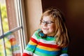 Adorable little preschool girl with eyeglasses sitting by the window. Thoughtful child looking out. Lonely kid. Royalty Free Stock Photo