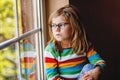 Adorable little preschool girl with eyeglasses sitting by the window. Thoughtful child looking out. Lonely kid. Royalty Free Stock Photo
