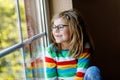 Adorable little preschool girl with eyeglasses sitting by the window. Thoughtful child looking out. Lonely kid. Royalty Free Stock Photo