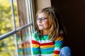 Adorable little preschool girl with eyeglasses sitting by the window. Thoughtful child looking out. Lonely kid. Royalty Free Stock Photo