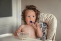 Adorable long haired toddler sitting in a baby high chair and eating porridge indoor. Royalty Free Stock Photo