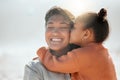 Adorable mixed race girl kisses her mother on the cheek and outs her arms around her while at the beach. Little daughter