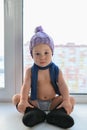 Adorable one year old baby boy lonely sitting on the windowsill weared in winter hat, shoes and scarf, indoors. Royalty Free Stock Photo
