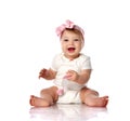 Adorable positive little baby girl in white body, hair bow decoration sitting on floor, smiling and holding toy in hand Royalty Free Stock Photo