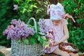 Adorable preschooler girl in pink plaid dress making lilac wreath in spring sunny garden
