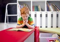 Adorable red-haired little boy, reading book in the library. Bookshelves. Student in preschool reading a magazine book