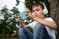 Adorable schoolboy wearing wireless headphones, playing with mobile phone, resting on the green grass of urban park Royalty Free Stock Photo
