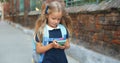 Adorable schoolgirl using modern smartphone while walking. Pretty kid with long blond hair and backpack going to school