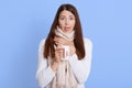 Adorable sick female drinking hot beverage and showing tongue, keeping hand on her neck wrapped in scarf, wearing white sweater, Royalty Free Stock Photo