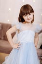 Adorable smiling little girl in festive princess blue dress is looking in camera in cozy room decorated lighting