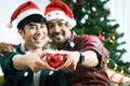 Adorable smiling young LGBT couple holding red heart and giving it to the camera, Asian gay male lover celebrating and sharing