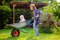 Adorable toddler boy having fun in a wheelbarrow pushing by mum in domestic garden, on warm sunny day Royalty Free Stock Photo