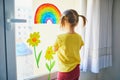 Adorable toddler girl attaching drawing of rainbow to window glass as sign of hope Royalty Free Stock Photo