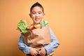 Adorable toddler holding paper bag with food standing over isolated yellow background