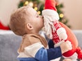 Adorable toddler kissing santa claus doll sitting on sofa by christmas tree at home