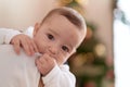 Adorable toddler sucking fingers standing by christmas tree on mother arms at home