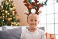 Adorable toddler wearing reindeer ears standing by christmas tree at home
