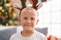 Adorable toddler wearing reindeer ears standing by christmas tree at home