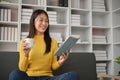 Adorable young Asian girl drinking a cup of coffee while reading book on sofa Royalty Free Stock Photo