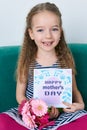 Adorable young girl sitting on a couch, holding bouquet of pink gerberas and Mother`s day card. Happy Mother`s Day Royalty Free Stock Photo