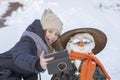 Adorable young girl is taking pictures of selfie with a snowman in beautiful winter park. Winter activities for children.