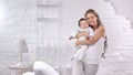 Adorable young mother hugging cute baby posing at white room cosiness interior looking at camera