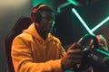 Adult African-American man in yellow hoodie and big black headphones racing on car simulator in gaming cafe. Royalty Free Stock Photo