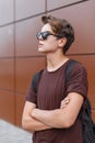 American young hipster man with backpack with trendy hairstyle in stylish sunglasses in fashionable brown t-shirt poses Royalty Free Stock Photo