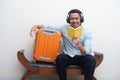 Adult Asian man sitting with travelling suitcase beside while listening music and read a book