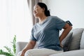 Adult Asian woman is sitting on the sofa and holding her lower back suffering from injured back. Health care and back pain concept Royalty Free Stock Photo