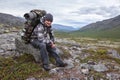 Adult Caucasian mountaineer resting on stone with heavy backpack on his back, man hiking in mountains, looking at camera, Royalty Free Stock Photo