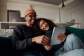 Trendy adult couple, multi-ethnic, laughing at video on tablet on sofa in modern apartment