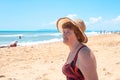 An adult elderly woman in a hat and a swimsuit on the sandy coast looks at the sea. Travel and tourism retired