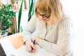 Adult focused blonde woman in glasses using laptop, writing notes in workplace at home. Remote training, learning. Female watching Royalty Free Stock Photo