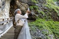 An adult gray-haired woman stands on the stairs in the Liechtensteinklamm gorge and shoots a video on her mobile phone