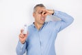 Adult handsome man with grey hair wearing blue shirt closing his nose smelling unpleasant perfume, standing on white