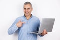 Adult handsome man with grey hair wearing blue shirt points with finger at his laptop standing on isolated white Royalty Free Stock Photo