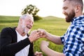 An adult hipster son with senior father making fist bump in nature at sunset.