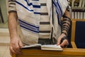 An adult jewish man praying with a tefillin on his arm, holding a bible book, while reading a pray